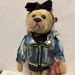 May include: A plush teddy bear wearing a denim jacket with a black zipper and black shorts. The bear has a black bow on its head and a tag attached to its right ear. The bear is a collectible item.