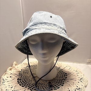 May include: A light blue denim bucket hat with a dark blue chin strap. The hat is displayed on a white mannequin head, resting on a cream-colored, lace doily.