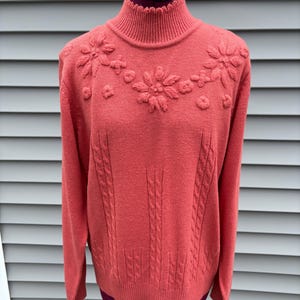 Puede incluir: Un suéter de color coral con cuello alto y con volantes, y mangas largas. El suéter presenta un diseño floral en el pecho y detalles de punto trenzado en la parte delantera. El suéter se muestra en un maniquí.