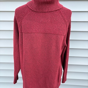May include: A burgundy turtleneck sweater with a textured knit design. The sweater features a ribbed turtleneck, a textured panel across the chest, and long sleeves. The bottom hem and cuffs are ribbed. The sweater is displayed on a mannequin.