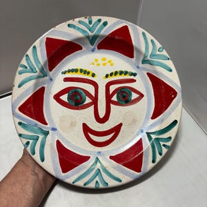 May include: A round ceramic plate featuring a hand-painted sun face design. The central face has red outlines for eyes, nose, and a smiling mouth, with green pupils. The sun rays are alternating red triangles and teal leaf-like patterns.