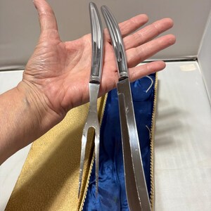 May include: A set of stainless steel carving tools, including a fork and a knife, presented in a gold and blue lined case. The tools have a sleek, modern design with curved handles.