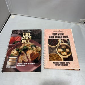 May include: Two spiral-bound cookbooks with the titles "Stack-Cooked Meals" and "Light & Easy Stack-Cooked Meals" by Tupperware. The covers feature images of prepared food. The first book has a dark cover, the second a light pink cover.