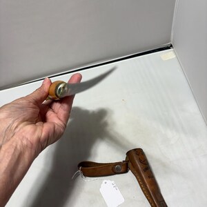 May include: A fixed-blade knife with a light-colored wooden handle and a metallic bolster, accompanied by a brown leather sheath featuring decorative stitching and a snap closure, with a small white price tag attached to the sheath.