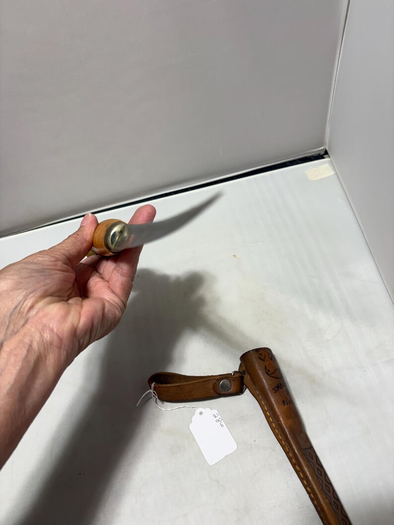 May include: A fixed-blade knife with a light-colored wooden handle and a metallic bolster, accompanied by a brown leather sheath featuring decorative stitching and a snap closure, with a small white price tag attached to the sheath.