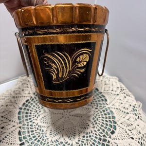 May include: A copper-colored metal bucket with a black panel featuring gold floral designs. The bucket has a scalloped rim and small metal handles. The bucket is sitting on a white lace doily.