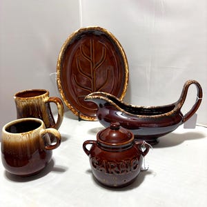 Pode incluir: Uma coleção de utensílios de cozinha de cerâmica marrom, incluindo duas canecas, uma travessa com um desenho de folha, uma molheira e um pequeno pote com tampa com a etiqueta "GARLIC". As peças têm um acabamento brilhante e um esquema de cores de dois tons.