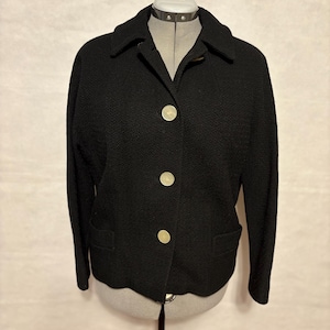 May include: A black jacket with a textured fabric, displayed on a mannequin. The jacket features a collared neckline, three large, round, cream-colored buttons, and two front pockets. The jacket's design is classic and tailored.