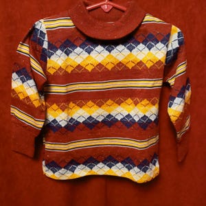 May include: A red, yellow, white, and blue striped sweater with a diamond pattern. The sweater has a mock neck and long sleeves.
