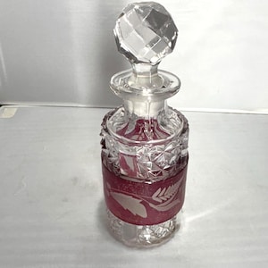 May include: A clear glass perfume bottle with a faceted stopper. The bottle has a red band with a leaf design. The bottle is decorated with cut glass patterns and is likely vintage.
