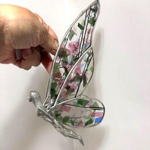 May include: A silver-toned fairy figurine with stained glass wings. The wings feature a mosaic of pink, green, and blue glass pieces, outlined with black lines. The fairy is held by a hand against a white background.