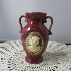 May include: A small, burgundy ceramic vase with a cameo of a woman in a white oval frame. The vase has a crackled glaze and gold accents.