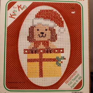May include: Counted cross stitch kit for kids featuring a dog wearing a Santa hat and holding a gift. The kit is labelled "Beginner Kid Tested Ages 6-96".