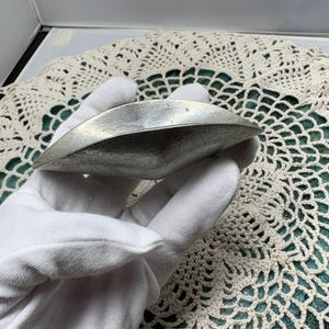 May include: A silver-colored, leaf-shaped dish. The dish has a smooth, reflective surface and is held by a gloved hand. The background features a cream-colored, crocheted doily on a green surface.