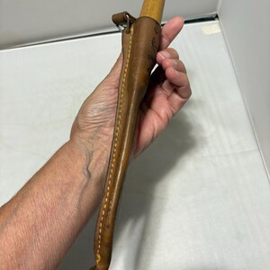 May include: A light brown leather sheath containing a wooden handled tool. The sheath features visible yellow stitching along the edge and a small metal loop attachment at the top. The wooden handle has a rounded, tapered end.