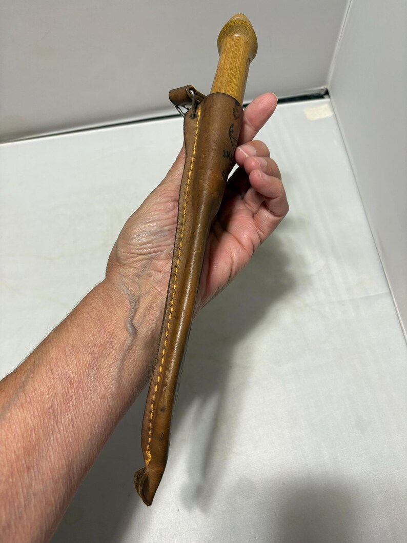 May include: A light brown leather sheath containing a wooden handled tool. The sheath features visible yellow stitching along the edge and a small metal loop attachment at the top. The wooden handle has a rounded, tapered end.