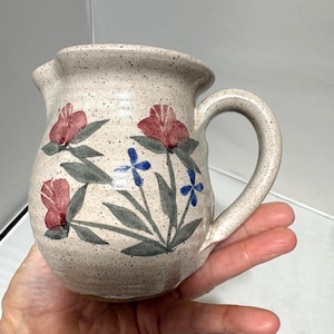 Puede incluir: Una jarra de cerámica moteada con asa y boquilla. La jarra está decorada con diseños florales rojos y azules y hojas verdes. La jarra se sostiene en una mano.