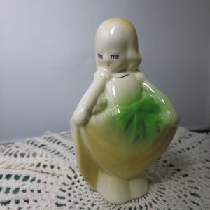 May include: A ceramic figurine of a girl with a green dress and a bow. The figurine is painted in shades of yellow and green.