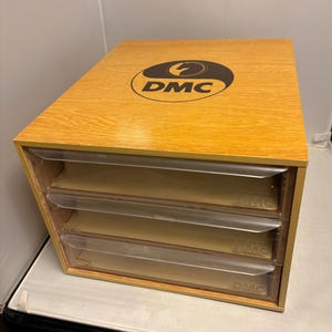 Puede incluir: Una caja de almacenamiento de madera con tres cajones de plástico transparente. La parte superior de la caja tiene un logotipo DMC negro y marrón. Los cajones tienen el logotipo DMC impreso. La caja es de madera clara.