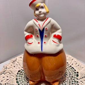 Puede incluir: Un tarro de galletas de cerámica vintage con forma de marinero. El tarro presenta una chaqueta blanca con detalles rojos y azules, un gorro de marinero y pantalones marrones. Un artículo de colección.