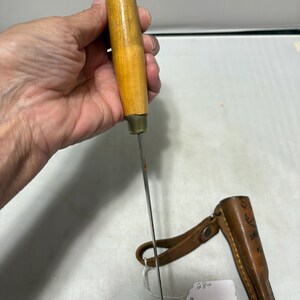 May include: A knife with a light brown wooden handle and a thin metal blade, accompanied by a brown leather sheath featuring a strap and a small white tag with the handwritten number 28.00 attached to the sheath by a white string.