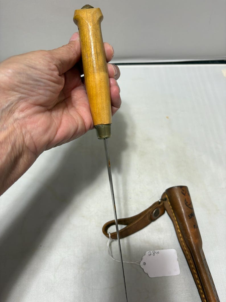 May include: A knife with a light brown wooden handle and a thin metal blade, accompanied by a brown leather sheath featuring a strap and a small white tag with the handwritten number 28.00 attached to the sheath by a white string.