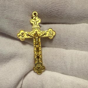 Puede incluir: Un colgante de cruz crucifijo dorado. La cruz presenta una representación detallada de Jesucristo y adornos decorativos en los brazos. El colgante está suspendido de un pequeño lazo en la parte superior.
