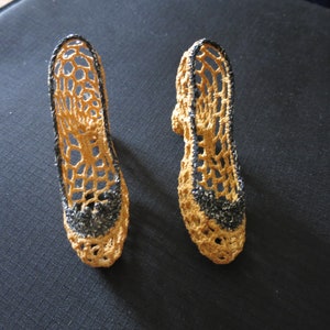 May include: A pair of handmade, open-weave, crochet shoes. The shoes are a light tan color with black trim around the edges and on the toe. The shoes are displayed on a dark surface.