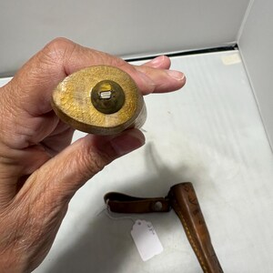 May include: A wooden handle with a brass-colored circular end cap featuring a small metal protrusion in the center. Below the handle, a brown leather sheath with embossed geometric patterns and a small white paper tag is visible.