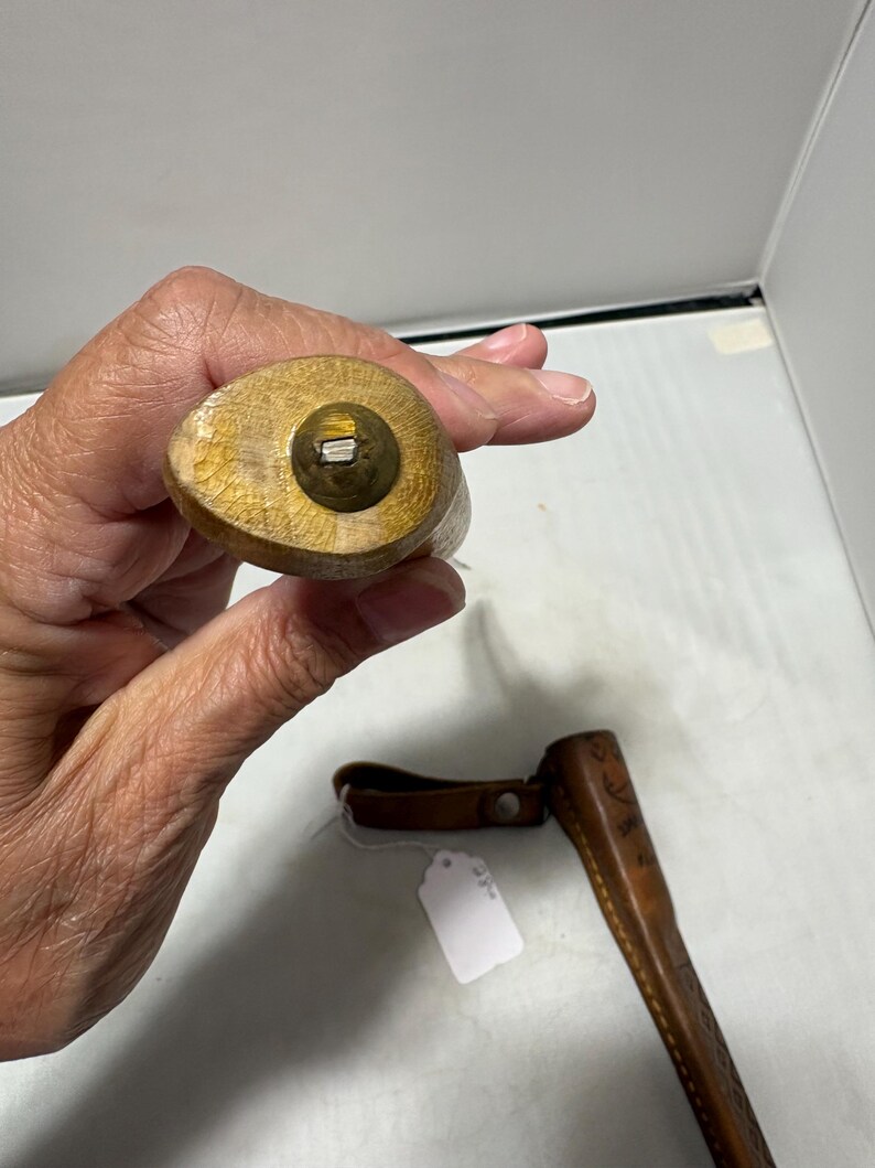 May include: A wooden handle with a brass-colored circular end cap featuring a small metal protrusion in the center. Below the handle, a brown leather sheath with embossed geometric patterns and a small white paper tag is visible.