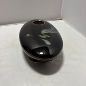 May include: A smooth, oval-shaped ceramic vessel with a glossy dark brown finish. The top surface features a subtle, light-colored abstract brushstroke pattern and a small circular opening with a thin rim. The object rests on three short feet.