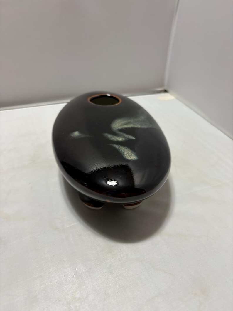 May include: A smooth, oval-shaped ceramic vessel with a glossy dark brown finish. The top surface features a subtle, light-colored abstract brushstroke pattern and a small circular opening with a thin rim. The object rests on three short feet.