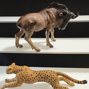 May include: A brown wildebeest figurine with a black mane and horns stands above a running cheetah figurine. The cheetah is tan with black spots. Both figurines are on a white surface.