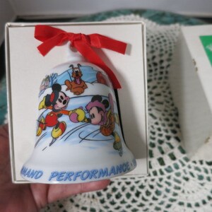 Puede incluir: Una campana de porcelana blanca con Mickey Mouse, Minnie Mouse y Pluto patinando sobre hielo. La campana tiene una cinta roja y las palabras "and Performance" en la parte inferior.