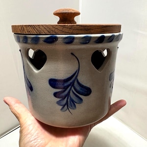 May include: A cylindrical ceramic crock with a light grey finish, featuring hand-painted dark blue leaf patterns and heart-shaped cutouts near the rim. The container is topped with a circular wooden lid that has a small, centred knob handle.