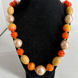 Puede incluir: Un collar vintage con una variedad de cuentas en tonos naranja, beige y crema. El collar presenta cuentas redondas, ovaladas y geométricas, ensartadas con detalles dorados. El collar se exhibe en un busto de joyería negro.
