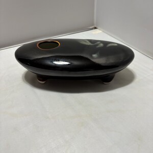 May include: A low, oval-shaped black ceramic vessel with a glossy finish and a single circular opening on the top surface. The piece is supported by three small, short feet and features a subtle, light-colored abstract glaze accent on one side.