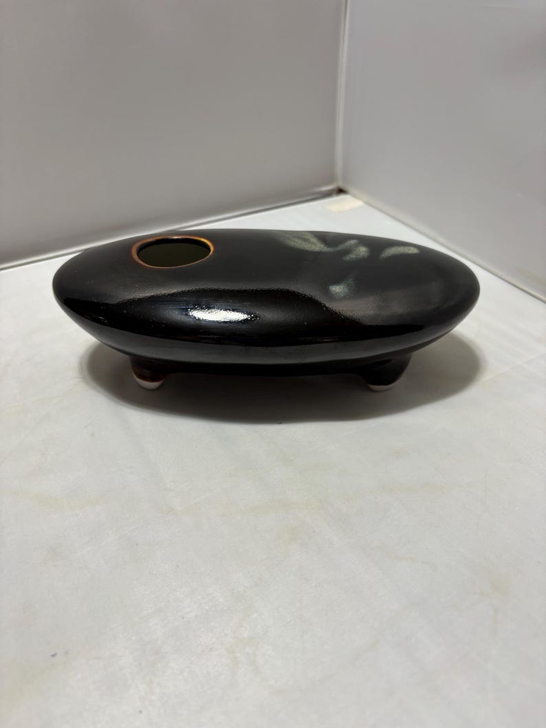 May include: A low, oval-shaped black ceramic vessel with a glossy finish and a single circular opening on the top surface. The piece is supported by three small, short feet and features a subtle, light-colored abstract glaze accent on one side.
