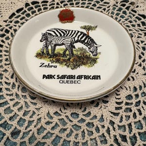 Puede incluir: Plato de cerámica blanco con una ilustración de cebra en blanco y negro. El plato tiene un borde dorado y el texto "Zebra Parc Safari Africain Quebec" impreso en él.