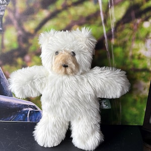 Peut inclure: Un ours en peluche avec une fourrure blanche longue et hirsute et un museau beige clair contrastant. L'ours a des yeux ronds foncés et un petit nez noir. Une étiquette en tissu vert est fixée sur le côté avec le texte Vermont Teddy Bear.