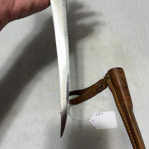 May include: A fixed-blade knife with a silver-toned metal blade, a brass-colored bolster, and a light-colored wooden handle. Beside the knife is a brown leather sheath with yellow stitching and a small white tag labeled with the number 28.