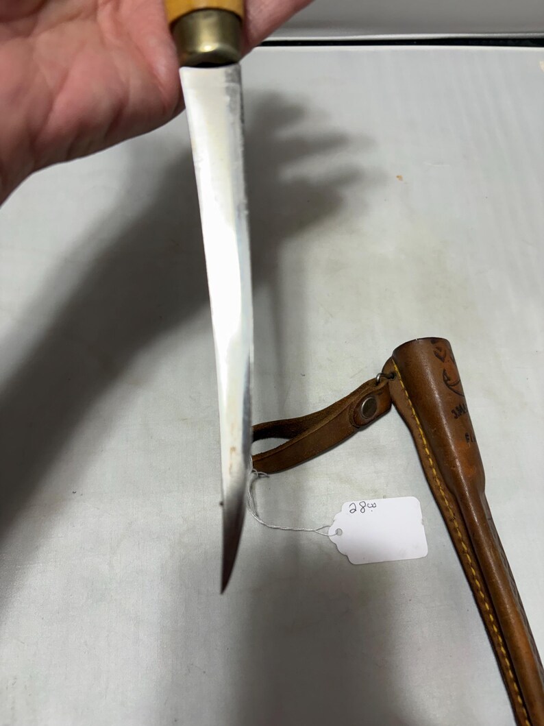 May include: A fixed-blade knife with a silver-toned metal blade, a brass-colored bolster, and a light-colored wooden handle. Beside the knife is a brown leather sheath with yellow stitching and a small white tag labeled with the number 28.
