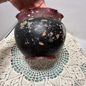 May include: A small, round, antique black pot with a ruffled, burgundy rim. The pot has a distressed appearance with visible paint chipping, revealing hints of orange and green. The base is also burgundy. The pot is sitting on a cream-colored, lace doily.