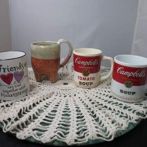 Może przedstawiać: Cztery ceramiczne kubki na białej koronkowej serwetce. Jeden kubek ma czerwony, biały i niebieski wzór serca z tekstem "Friends are tied together with heartstrings". Dwa kubki mają logo Campbell's Soup i tekst "Tomato Soup". Jeden kubek jest brązowo-biały w kropki z trzema nóżkami.