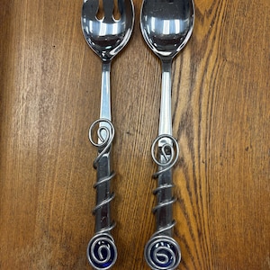 May include: A pair of stainless steel salad serving utensils consisting of a fork and a spoon. Each handle features a decorative silver-toned metal wire wrapped in a spiral pattern, terminating in a circular dark blue glass accent at the base.