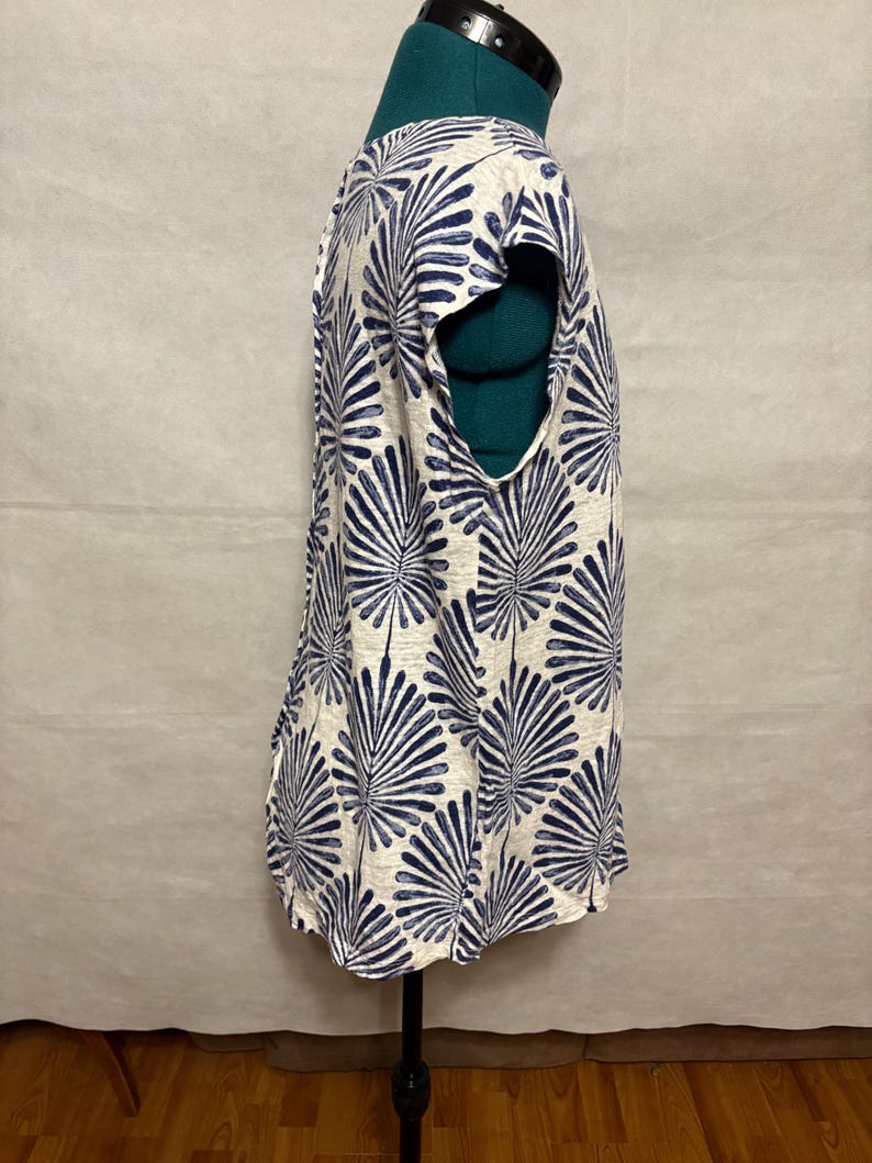 K&ouml;nnte beinhalten: Ein &auml;rmelloses Oberteil mit wei&szlig;em Hintergrund und einem sich wiederholenden blauen Bl&auml;ttermuster. Das Kleidungsst&uuml;ck wird auf einer Schaufensterpuppe pr&auml;sentiert, um sein Design hervorzuheben. Der Stoff wirkt leicht und f&uuml;r warmes Wetter geeignet.