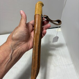 May include: A knife with a light-colored wooden handle encased in a brown leather sheath. The sheath features visible stitching along the edge and includes a leather belt loop attached with a metal ring. A small white price tag hangs from the loop.