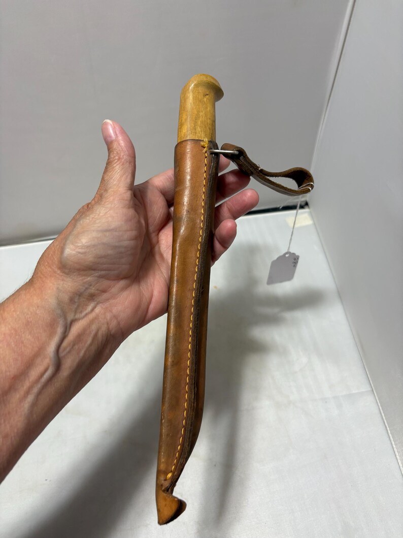 May include: A knife with a light-colored wooden handle encased in a brown leather sheath. The sheath features visible stitching along the edge and includes a leather belt loop attached with a metal ring. A small white price tag hangs from the loop.