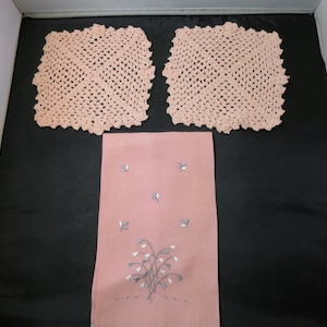 May include: Two pink crocheted square doilies and a pink linen tea towel with embroidered white flowers.