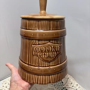 Pode incluir: Pote de biscoitos de cerâmica marrom com tampa e alça. O pote tem formato de barril com linhas verticais e faixas horizontais. As palavras "COOKIE CHURN" estão em relevo na frente. O pote é segurado por uma mão.